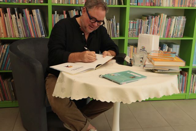 autor podpisuje książkę przy stoliku, w tle regały z książkami