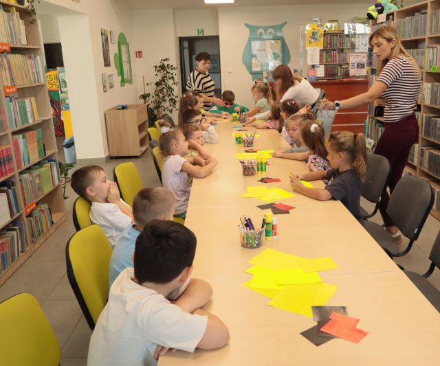 przedszkolaki siedzą przy połączonych stołach, na stołąch żółte kartki i nożyczki