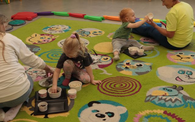 małe dzieci siedzą na dywanie ukłądają drobne przedmioty w papierowych kubeczkach