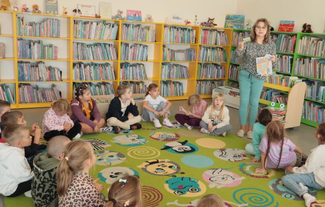 dzieci siedzą w okręgu na kolorowym dywanie, w tle regały z książkami