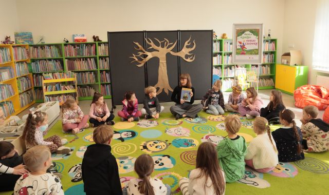 przedszkolaki i bibliotekarka siedzą na kolorowym dywanie, w tle plansza z dużym drzewem i regały z książkami