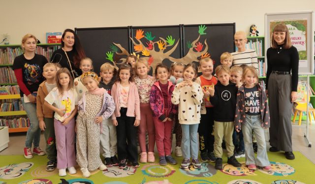 przedszkolaki, bibliotekarka i panie nauczycielki pozują do zdjęcia, w tle plansza z dużym drzewem i regały z książkami