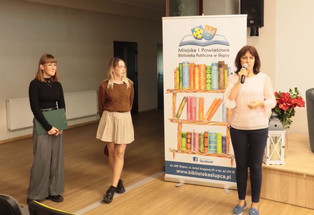 na zdjęciu kobieta z mikrofonem oraz dwie kobiety, w tle baner z logo i nazwą biblioteki