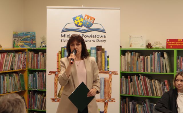 dyrektorka biblioteki stoi z mikrofonem na tle baneru z logo biblioteki