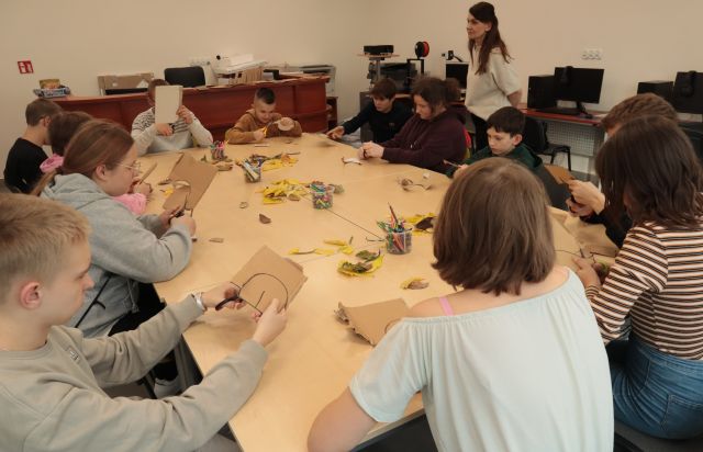 młodzież siedzi przy ustawionych połączonych w kwadrat stołach, na stołach pocięte kartony, kleje, nożyczki