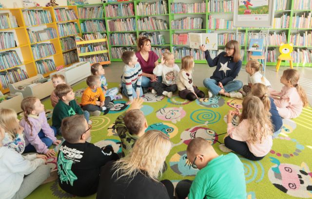 bibliotekarka z książką i przedszkolaki siedzą na kolorowym dywanie, w tle regały z książkami
