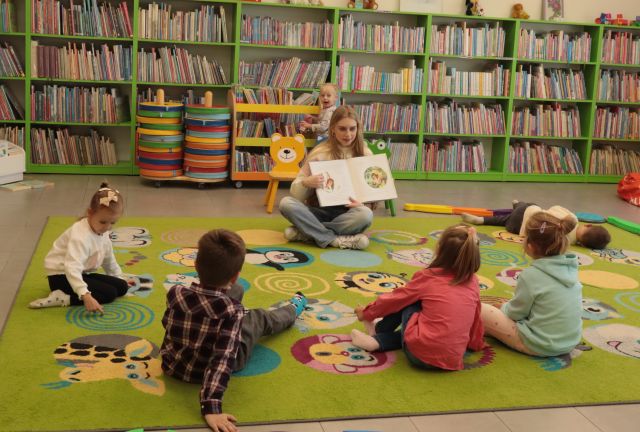 bibliotekarka z książką siedzi przed 5 małych dzieci siedzących na dywanie, w tle regały z książkami