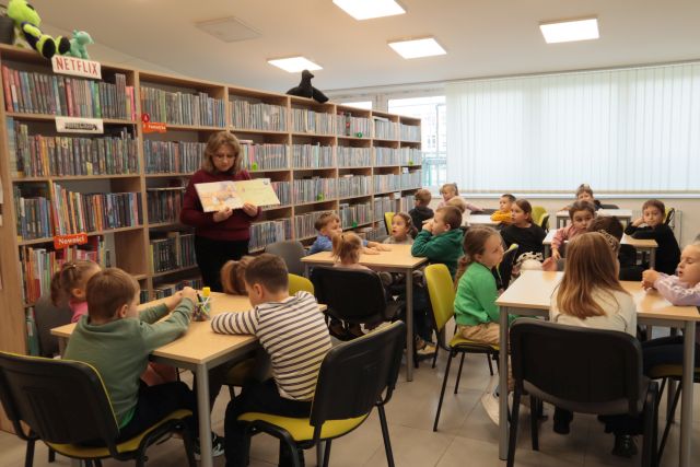 dziesi siedzą przy stolikach, przed nimi stoi bibliotekarka z książką