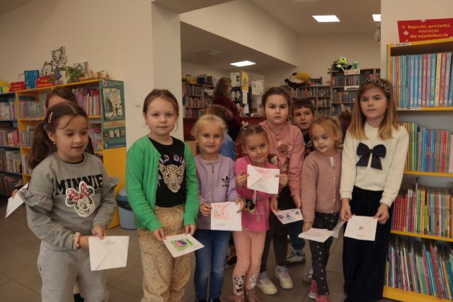 grupka przedszkolaków prezentuje swoje prace konkursowe, w tle regały z książkami