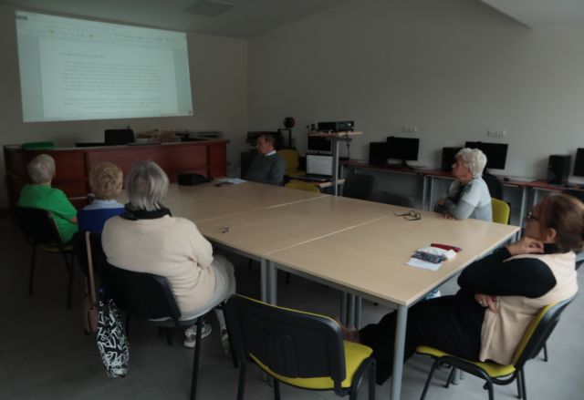 6 seniorów siedzi przy połączonych stołach i ogląda prezentację