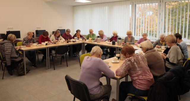 20 seniorek siedzi przy stołach ustawionych w literę u i pisze