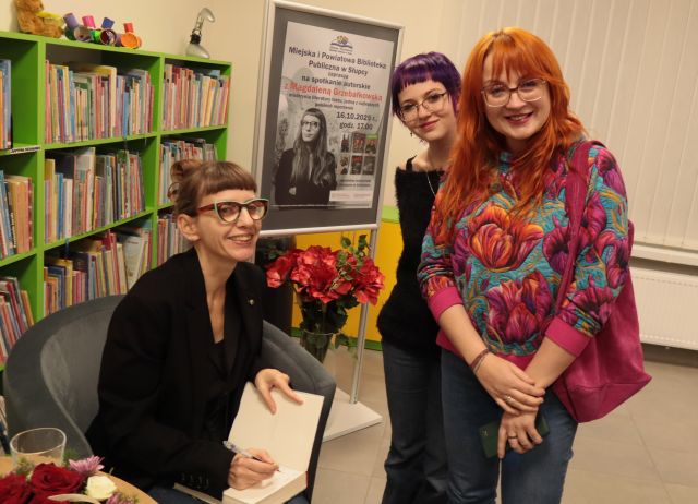autorka podpisuje książkę, obok niej stoją dwie kobiety