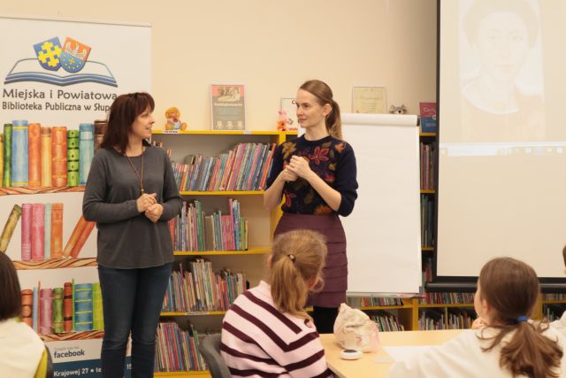 przed dziećmi siedzącymi przy stolikach stoi dyrektorka biblioteki i ilustratorka, w tle regały z książkami