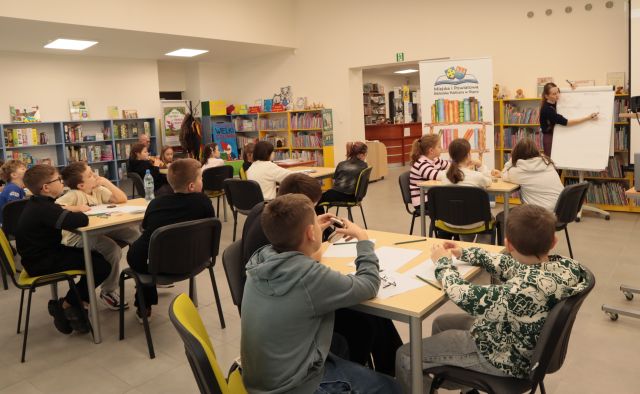 18 dzieci siedzi przy stolikach, na stolikach kartki, ołówki i kredki świecowe