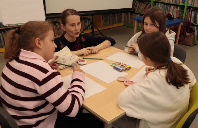 przy 3 dziewczynach siedzących przy stole z kartkami kuca ilustratorka