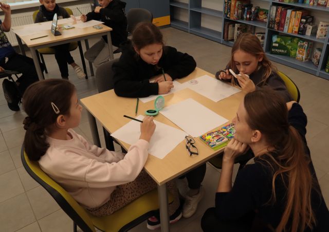 przy 3 dziewczynach siedzących przy stole z kartkami kuca ilustratorka