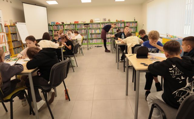 18 dzieci siedzi przy stołach, w tle ilustratorka i regały z książkami