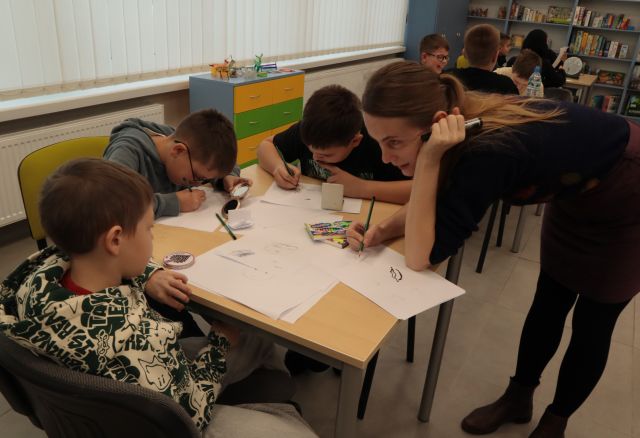 ilustratorka pochylona nad 3 rysujących dzieci