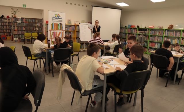 przy stolikach siedzi 18 dzieci rysujących na kartkach, w tle ilustratorka przy flipcharcie