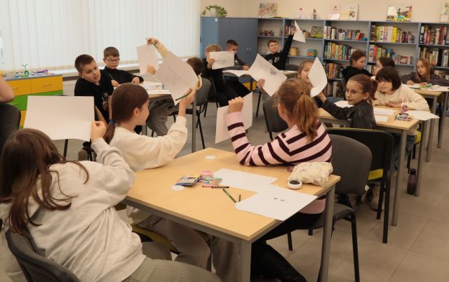 dzieci siedzące przy stolikach z podniesionymi do góry kartkami