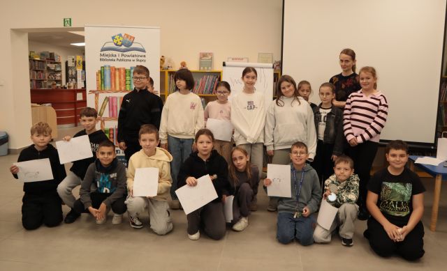 18 dzieci uczestniczących w warsztatach pozuje do zdjęcia z kartkami i ilustratorką