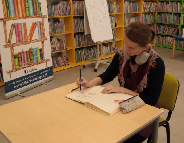 ilustratorka wpisuje się do kroniki biblioteki, w tle regały z książkami