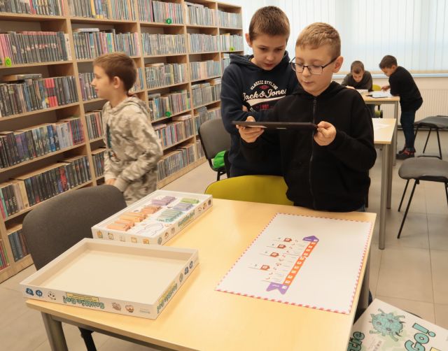 chłopiec tabletem robi zdjęcie planszy gry, w tle inni chłopcy i regały z książkami