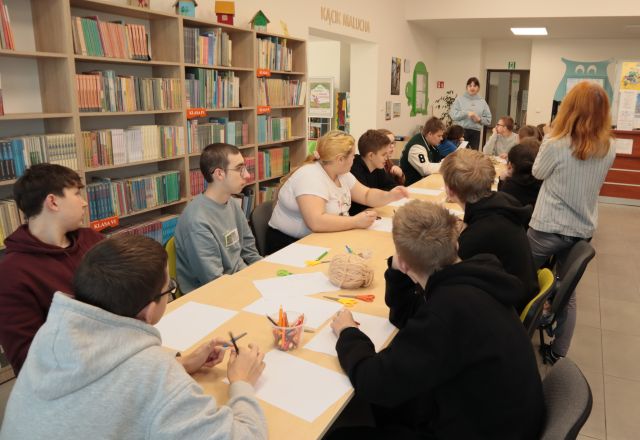 młodzież siedzi przy ustawionych w długim rzędzie stołach, w tle regały z książkami