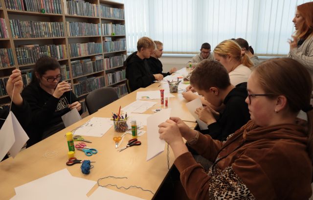 młodzież nawlekająca wełnę na igły, na stołach leżą kartki i nożyczki, w tle regały z książkami