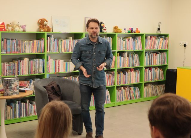 na zdjęciu pisarz stojący przed siedzącą młodzieżą, w tle regały z książkami