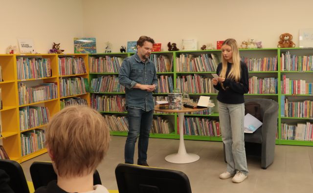 na środku sali stoi autor i dziewczynna czytająca z kartki, w tle regały z książkami