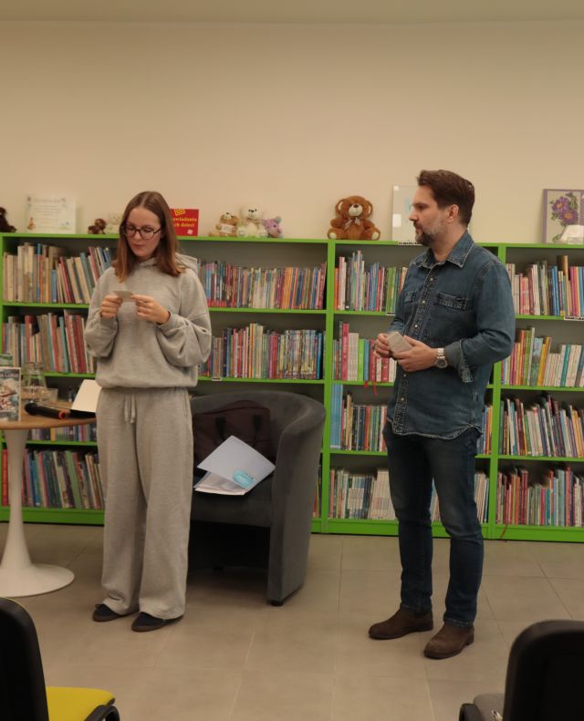na środku sali stoi autor i dziewczynna czytająca z kartki, w tle regały z książkami