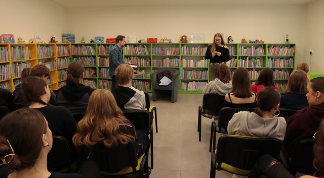 autor i czytająca dziewczyna stoją przed siedzącą na krzesłach młodzieżą, w tle regały z książkami