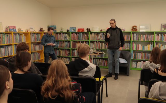 autor i czytający chłopak stoją przed siedzącą na krzesłach młodzieżą, w tle regały z książkami