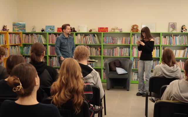 autor i czytająca dziewczyna stoją przed siedzącą na krzesłach młodzieżą, w tle regały z książkami