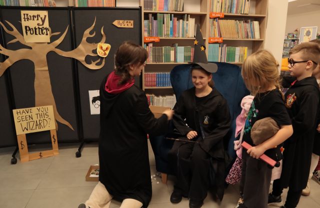 bibliotekarka w pelerynie klęczy przed dziewczynką w stroju Harry'ego Pottera siedzącą na fotelu, w tle inne dzieci i regały z książkami