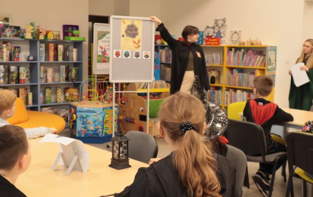 bibliotekarka w pelerynie stoi przded dziećmi w strojach z Harry'ego Pottera siedzących przy stołach, w tle regały z książkami