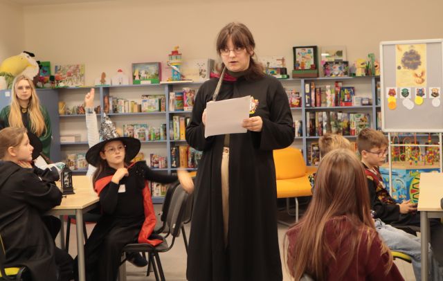 bibliotekarka w pelerynie z kartką zstoi przed siedzącymi przy krzesłach dziećmi w strojach z Harry'ego Pottera