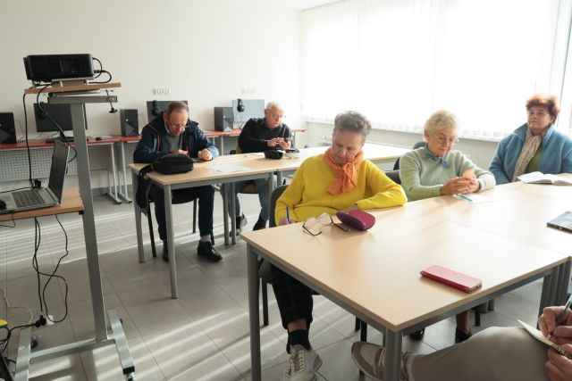 9 seniorów siedzi przy stołach, przed nimi rzutnik