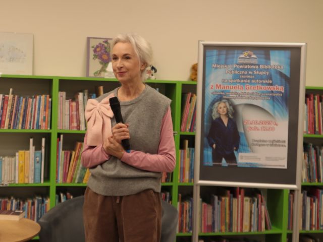 Manuela Gretkowska stoi z mikrofonem, z prawej strony plakat zapraszający na spotkanie, w tle regały z książkami
