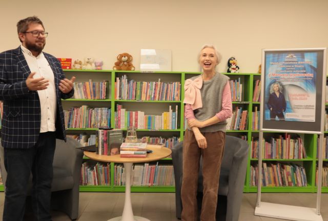 prowadzący i autorka stoją przy okrągłym stoliku, w tle regały z książkami
