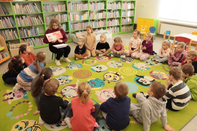 przedszkolaki siedzą na kolorowym dywanie, bibliotekarka siedzi na krzesełku, w tle regały z książkami