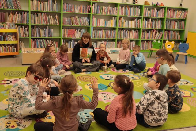 przedszkoalki i bibliotekarka z książką siedzą na kolorowym dywanie