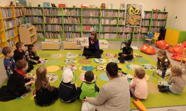 dzieci i bibliotekarka z książką siedzą na kolorowym dywanie, w tle regały z książkami