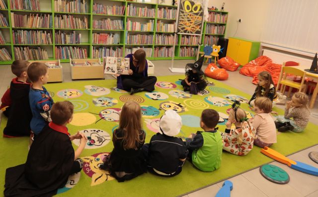 dzieci i bibliotekarka z książką siedzą na zielonym dywanie, w tle regały z książkami