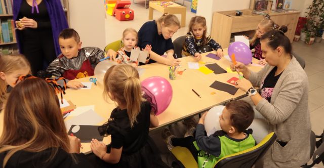 dzieci i rodzice siedzą przy stołach, rysują i wycinają, na stołąch leżą kartki i balony