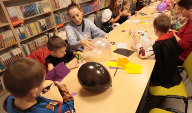 dzieci i rodzice siedzą przy stołach, rysują i wycinają, na stołach leżą kartki i balony