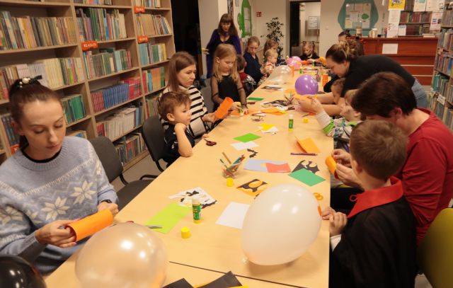 dzieci i rodzice siedzą przy stołach, malują i wycinają, na stołach leżą kartki, kredki, balony