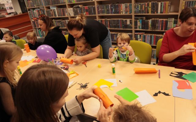 dzieci i rodzice siedzą przy stołach, malują i wycinają, na stołach leżą kartki, kredki, balony