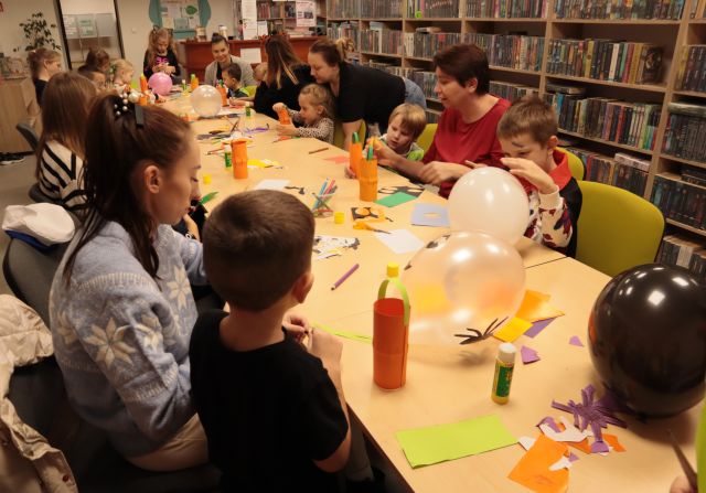 dzieci i rodzice siedzą przy stołach, malują i wycinają, na stołach leżą kartki, kredki, balony